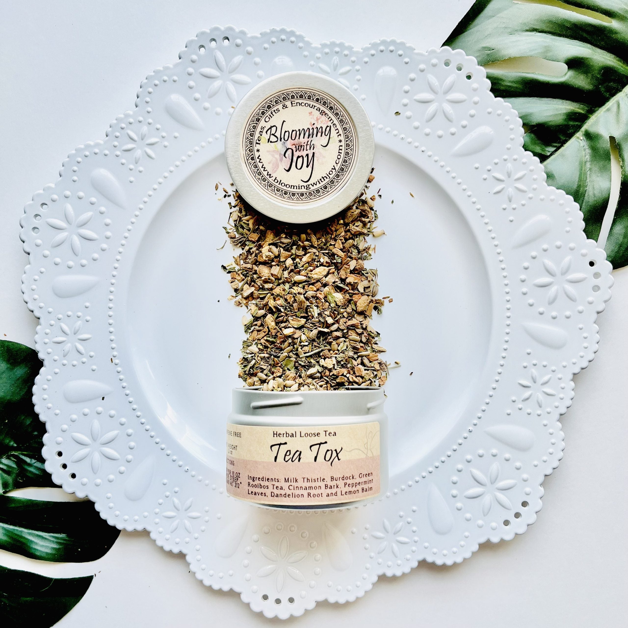 Blooming with Joy Tea Tox herbal loose leaf tea, showing dry herbs spilling from a small jar onto a white decorative plate with green leaves in the background.